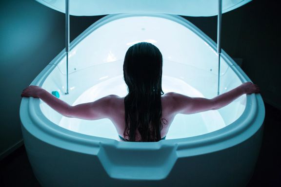 A person in a bathing suit sits facing away inside an illuminated floating tank.