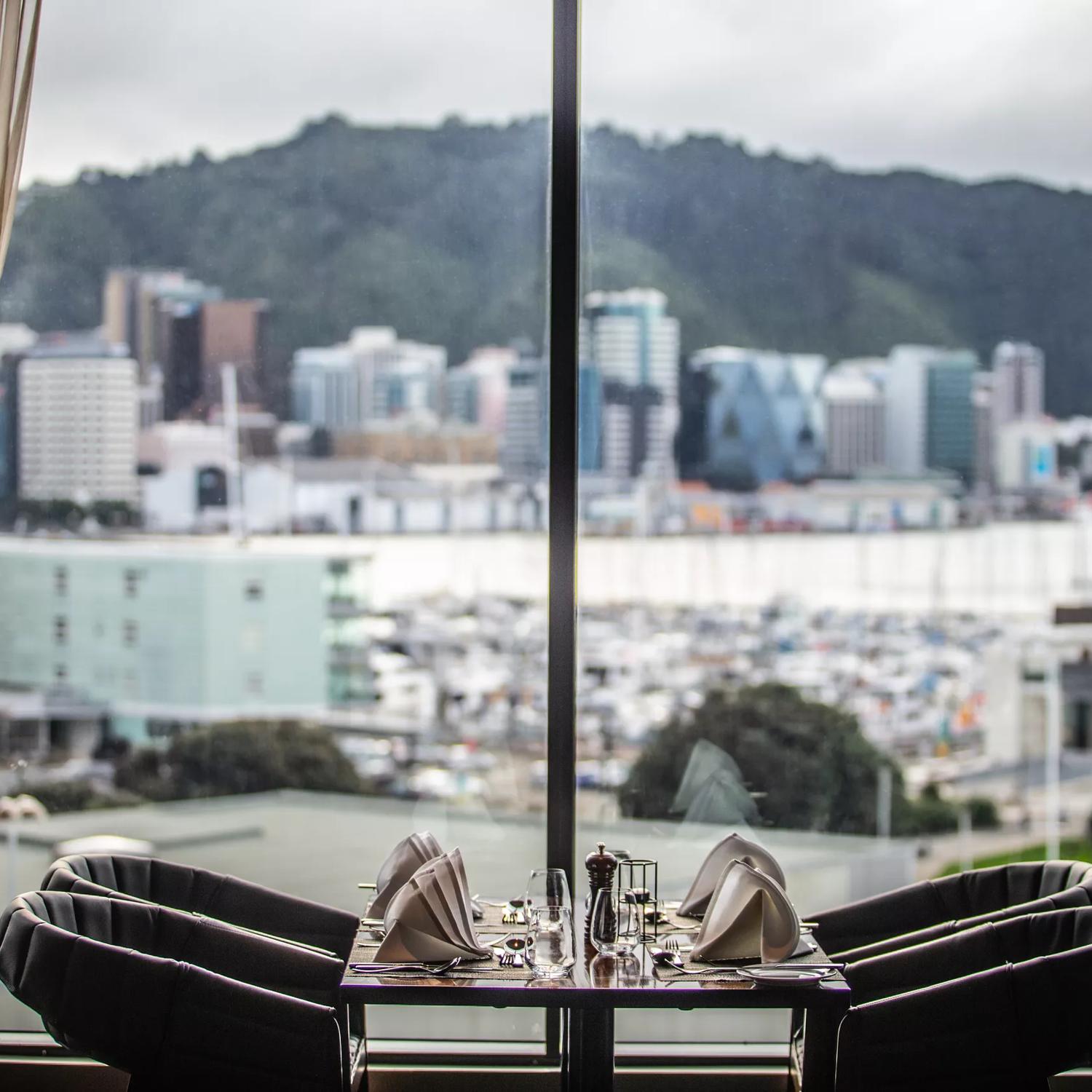 The view from the Copthorne Hotel dining room, 4 chairs sit at a table right next to a floor to ceiling window, overlooking the waterfront and Wellington harbour.