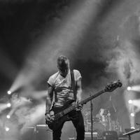Peter Hook stands on stage playing the bass guitar. Stage lights illuminate the background with smoke in the air, the drummer is behind him. The image is black and white.