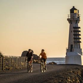 2 bikers on the Pencarrow Coast Road at sunset, with the lighthouse in the background.