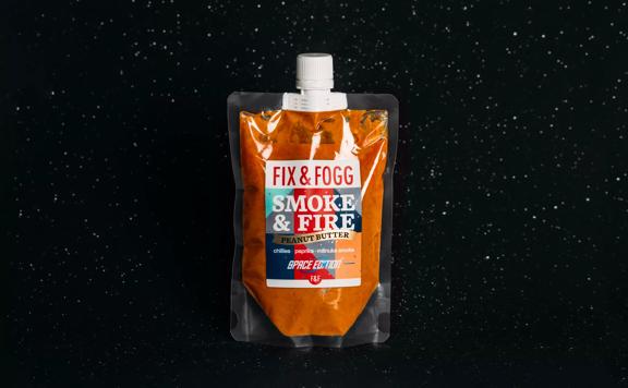 Fix & Fogg Smoke & Fire space pouch floats in space. The bright orange peanut butter is in a clear plastic pouch with a white plastic twist cap and a white, red, blue and grey label. 
