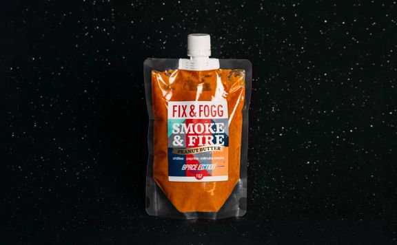 Fix & Fogg Smoke & Fire space pouch floats in space. The bright orange peanut butter is in a clear plastic pouch with a white plastic twist cap and a white, red, blue and grey label.