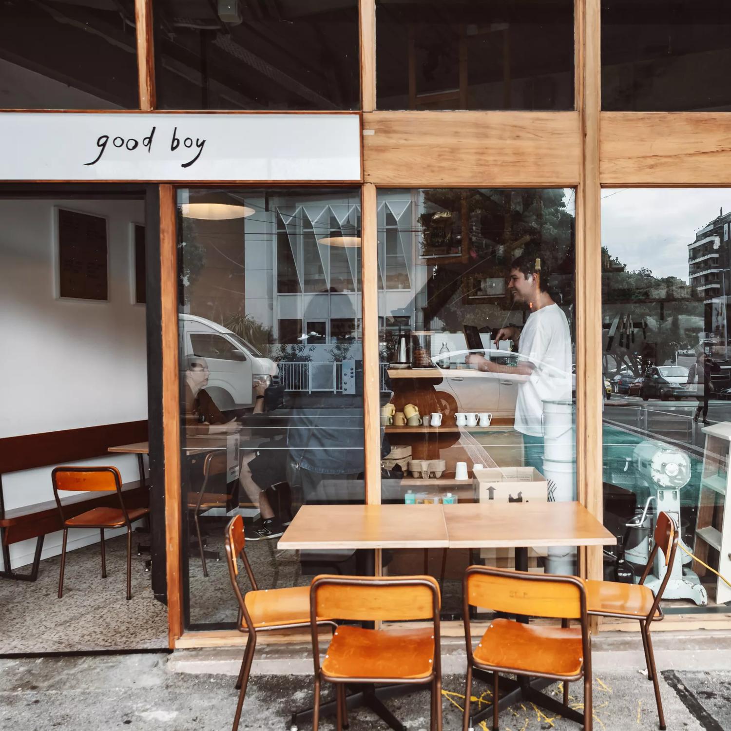 The exterior of Good Boy, a small sandwich shop and café in Newtown, Wellington. The front facade is made of large windows with wood framing and there is a small table outside with four chairs.