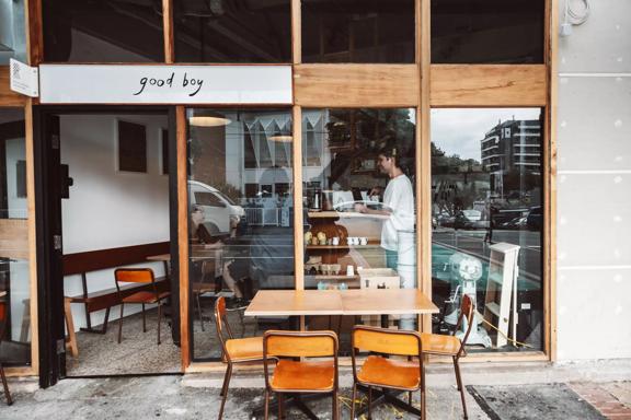 The exterior of Good Boy, a small sandwich shop and café in Newtown, Wellington. The front facade is made of large windows with wood framing and there is a small table outside with four chairs.