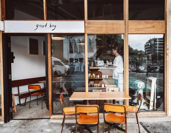 The exterior of Good Boy, a small sandwich shop and café in Newtown, Wellington. The front facade is made of large windows with wood framing and there is a small table outside with four chairs.