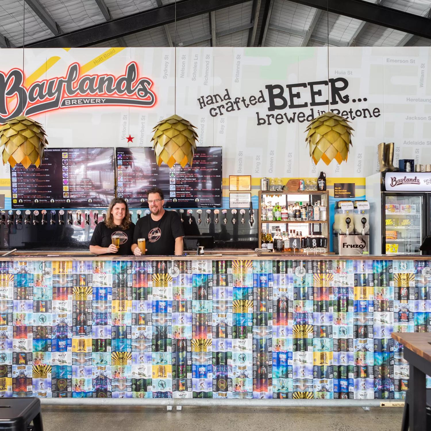 A wide look at the main bar at Baylands Brewery, with two bartenders posing in front of the beer taps, with a beer in hand.