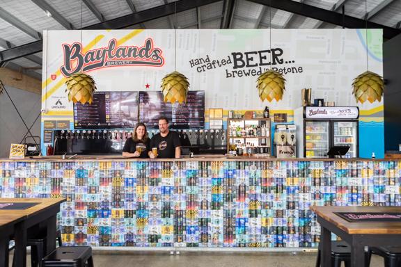 A wide look at the main bar at Baylands Brewery, with two bartenders posing in front of the beer taps, with a beer in hand.