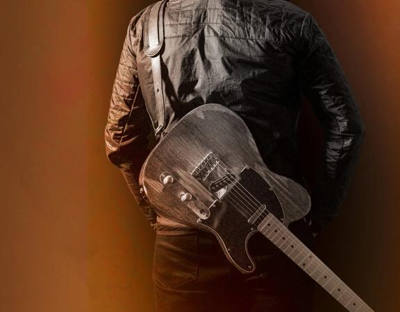 A person in a leather jacket with an electric guitar stepped to their back facing away on a brown-orange gradient background.