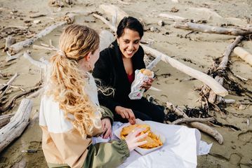 Two people sit on a driftwood-strewn beach sharing takeaway fish and chips from paper containers, with sandy shoreline and scattered logs in the background.