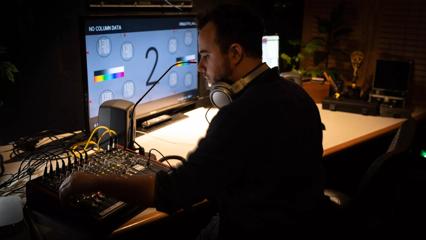 A person sits at a desk with headphones around their neck, working with a sound desk.