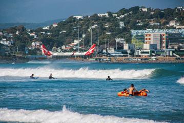 People surfing and swimming in the waves at Lyall Bay Beach with Wellington Airport visible in the distance.