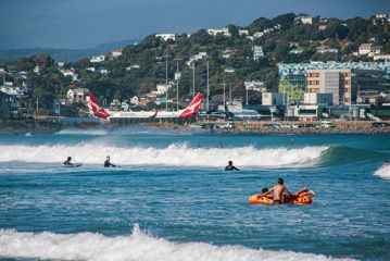 People surfing and swimming in the waves at Lyall Bay Beach with Wellington Airport visible in the distance.