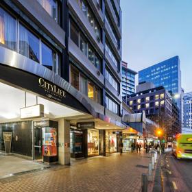 Exterior of CityLife Hotel, looking down Lambton Quay at dusk as commuters walk on the footpath and a bus goes by.