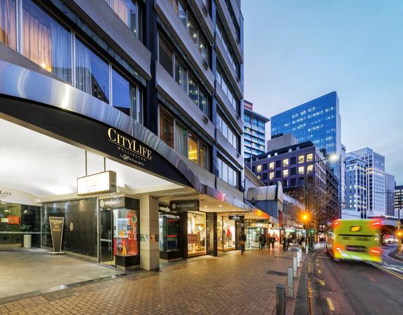 Exterior of CityLife Hotel, looking down Lambton Quay at dusk as commuters walk on the footpath and a bus goes by.
