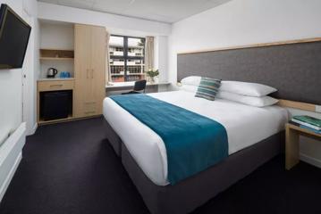 Room inside Atura Hotel, showing a double bed with blue blanket, desk, and kitchenette.