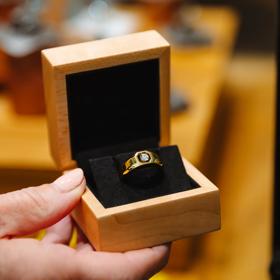 A hand holds a wooden ring box that has a Floeting diamond ring placed in it.