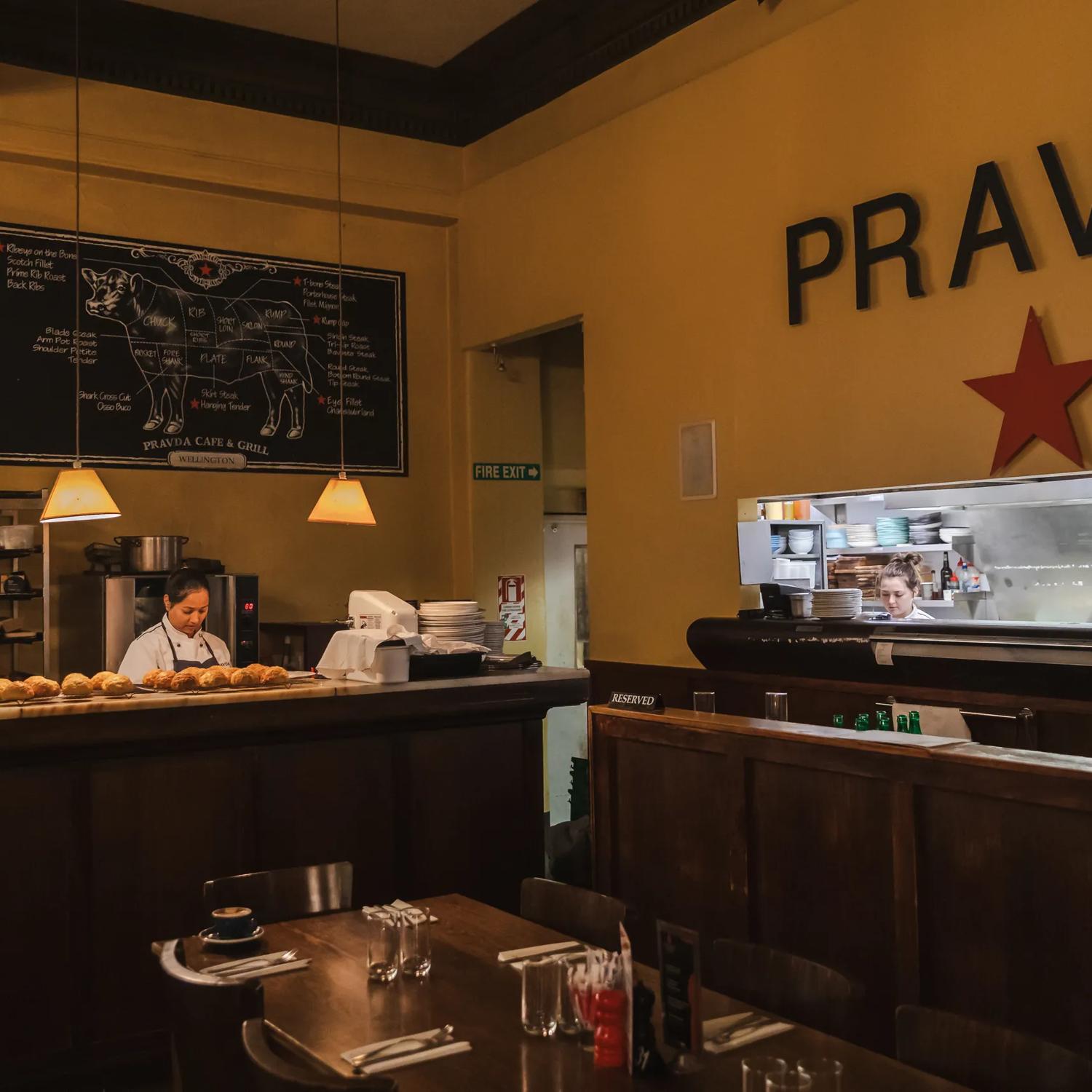 The interior Pravda Café and Grill is dimly lit with high ceilings and dark wood. A person sits in the corner booth table while a baker works behind the counter with freshly bakes cheese scones cooling on baking racks.