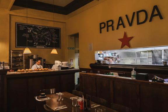 The interior Pravda Café and Grill is dimly lit with high ceilings and dark wood. A person sits in the corner booth table while a baker works behind the counter with freshly bakes cheese scones cooling on baking racks.