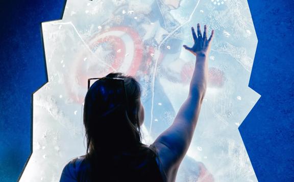 A person stands with their arm raised and touching a Captain America display at the Marvel: Earth's Mightiest Exhibition at Tākina Wellington Convention & Exhibition Centre.