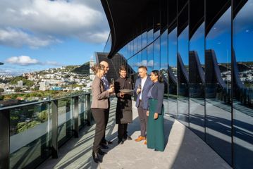Four people in business dress are served by a waiter wearing black. They stand on the balcony of Tākina, looking out over Wellington.