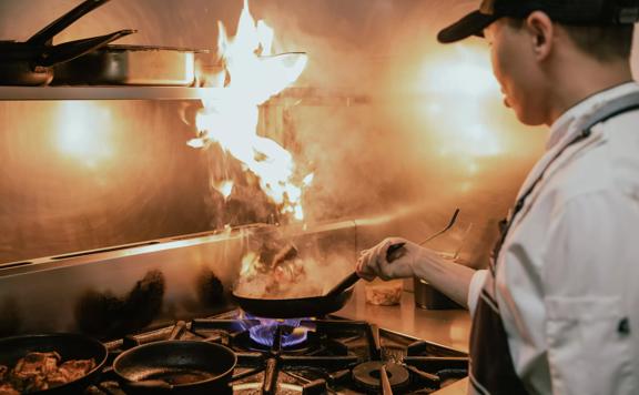 Close-up of chef in the kitchen at Apache, tossing up meat in a pan with fire coming off the oil.