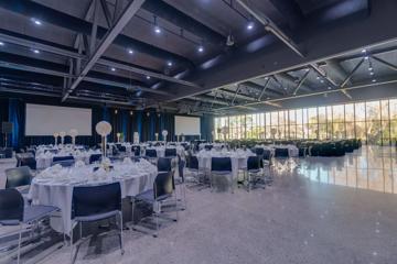 A large room inside the Lower Hutt Events Centre, with high ceilings, floor-to-ceiling windows, and roughly 50 tables, each set up with glasses and plates.