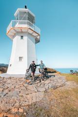 Two cyclists pose for a photo in front of a small white lighthouse.