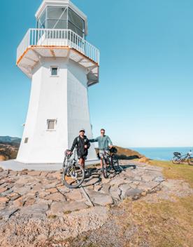 Two cyclists pose for a photo in front of a small white lighthouse.