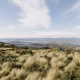 A vast landscape with tall dry grass in the foreground, rolling hills, houses along the coastline and a mountain range across the water.