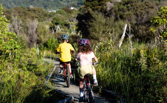 Two children biking away from the camera on the Wetland Loop trail in Wainuiomata Bike Park.