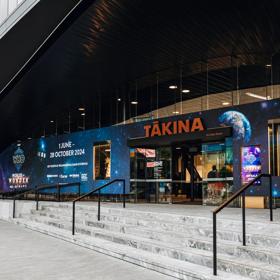 Exterior facade of the Takina building with large space-themed decals on the windows. There are stairs leading up to the entrance, with black handrails.
