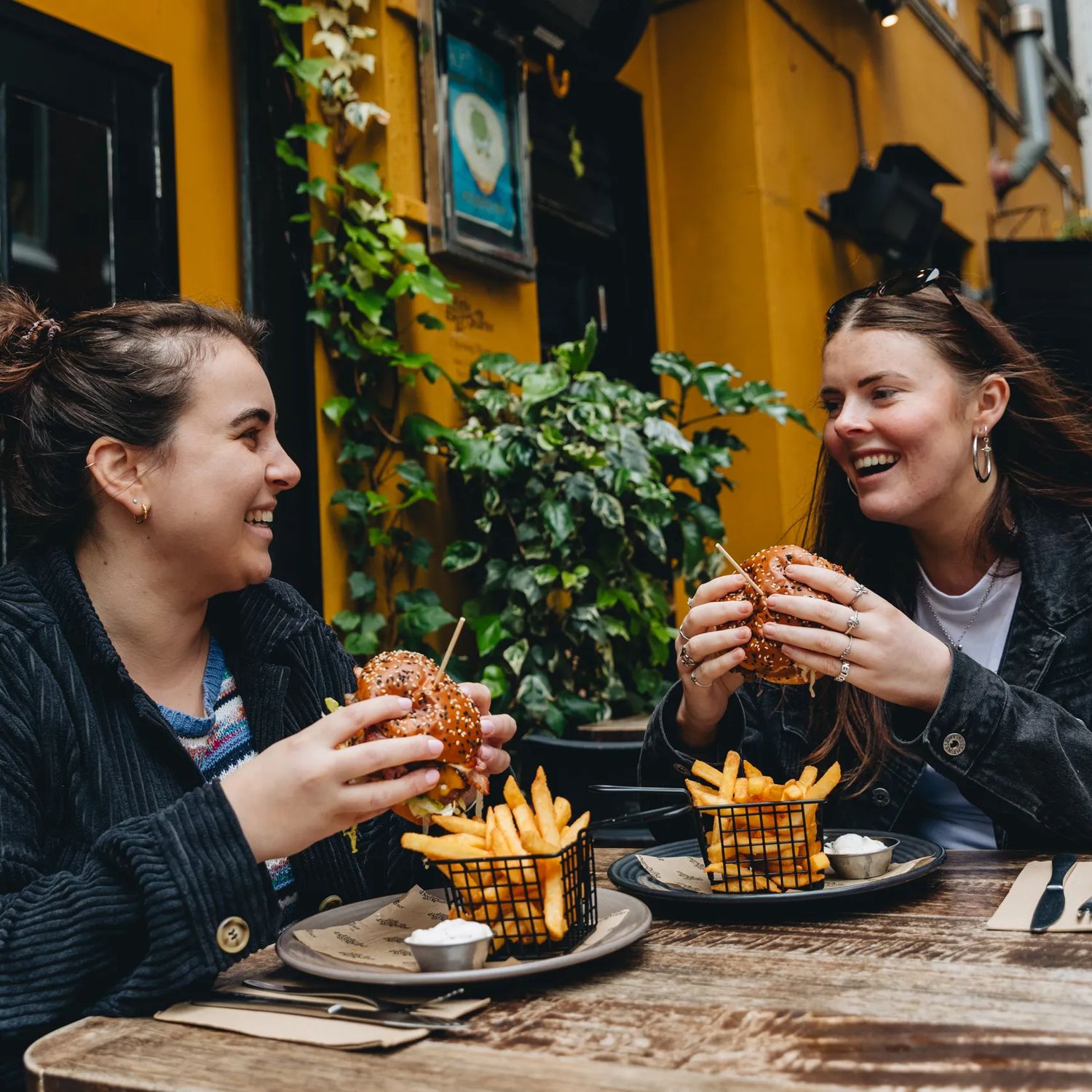 Two smiling people sitting outside at a wooden table, holding burgers