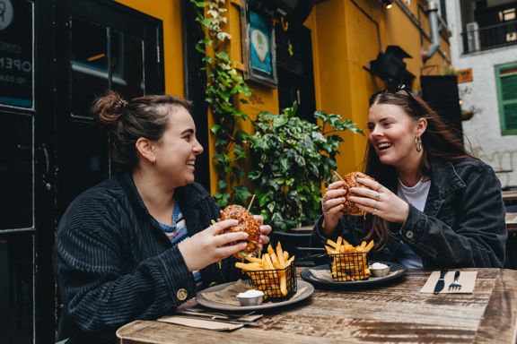 Two smiling people sitting outside at a wooden table, holding burgers