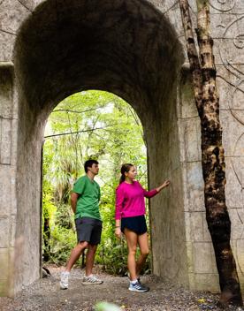 Two people walking through a large monument sitting between trees and bush on the Kaitoke regional park trail.