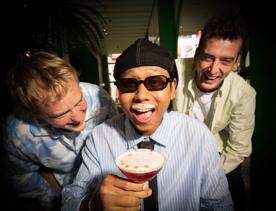 Three friends are laughing together. The one in the middle has a foam moustache from sipping a fancy cocktail from a martini glass.