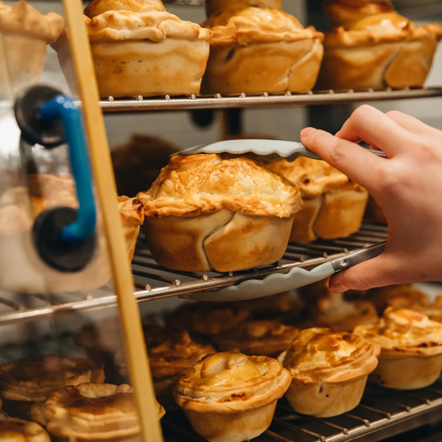 A person uses tongs to pick up a pie from a metal rack.