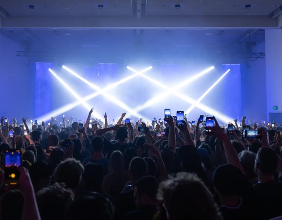 Large crowd at an indoor concert, with bright stage lights crossing above the audience and many attendees holding up phones to record the performance.