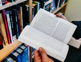 Hands holding a novel open with Te reo Māori books in the background.