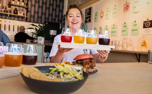 A smiling server carries a flight of craft beers to a table at Heyday Beer Co, a brewery located on upper Cuba Street in Te Aro, Wellington. 