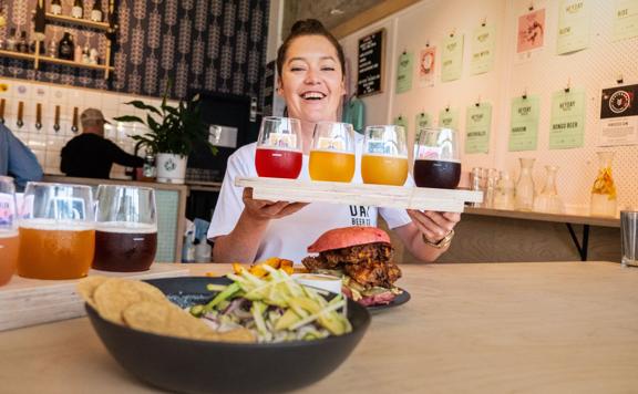 A smiling server carries a flight of craft beers to a table at Heyday Beer Co, a brewery located on upper Cuba Street in Te Aro, Wellington.