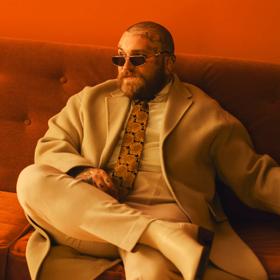 American singer-songwriter, Teddy Swims’ wearing a swanky outfit sitting on a couch in a room with red lighting. 