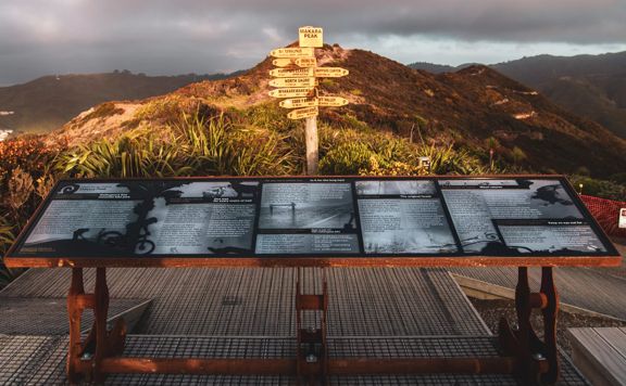 A large informational plaque at Mākara Peak Mountain Bike Park with a directional signpost centred behind it and a mountain range in the background.