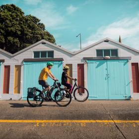 Two bicyclists ride together in front of the colourful boat sheds located in Oriental Bay in Wellington.