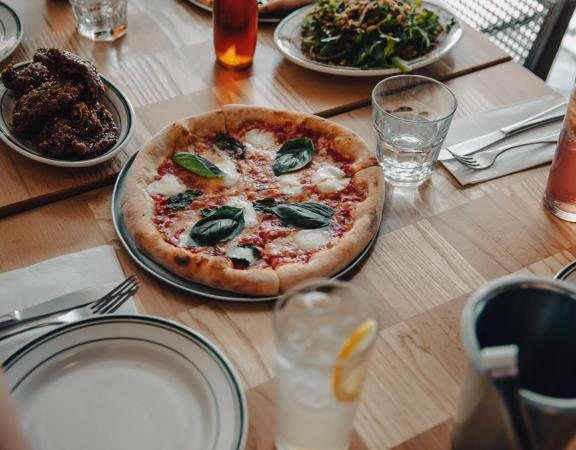 A pizza sits in the middle of a table at Scopa, with glasses, cutlery, and 2 side plates surrounding it.