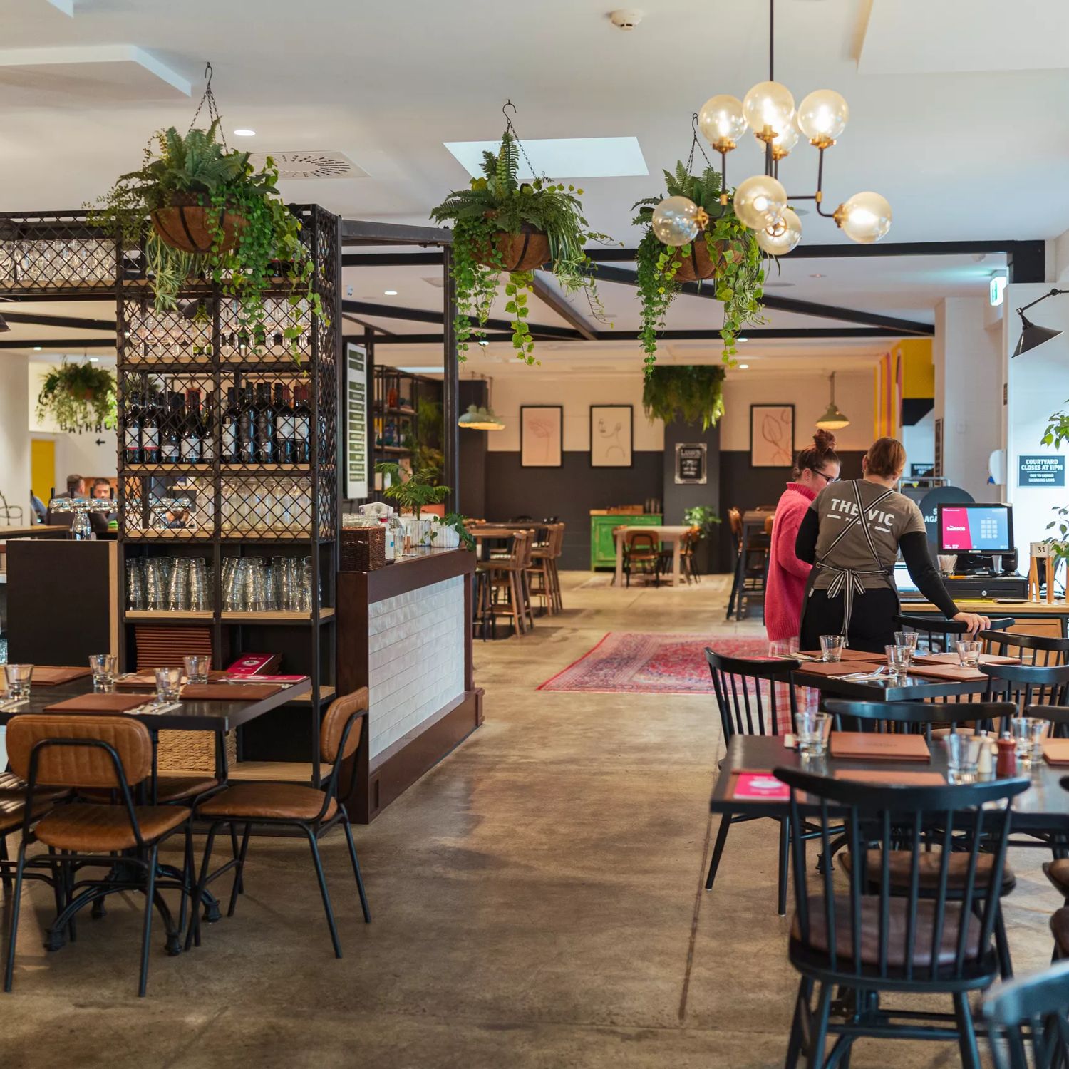 The interior of The Victoria Tavern located in Petone in Lower Hutt. It is a spacious, luminous restaurant with concrete floors, tables of varying sizes, chairs and two staff members standing at the front desk.