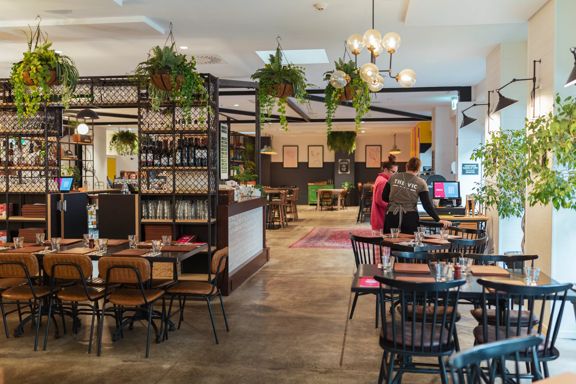 The interior of The Victoria Tavern located in Petone in Lower Hutt. It is a spacious, luminous restaurant with concrete floors, tables of varying sizes, chairs and two staff members standing at the front desk.
