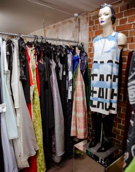 Many floor-length dresses hang on a rack in various colours and patterns. A mannequin with no arms stands on a box wearing a tunic with cloud print and the word ‘peace’.