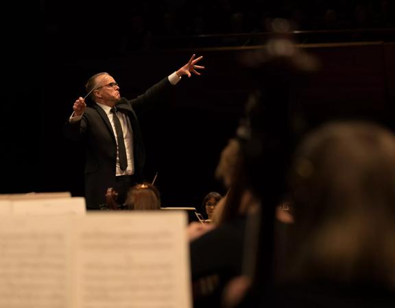 Image of Marc Taddei conducting Orchestra Wellington. His right hand has a baton in it while the other hand is outstretched. In the foreground, blurred music scores can be seen.