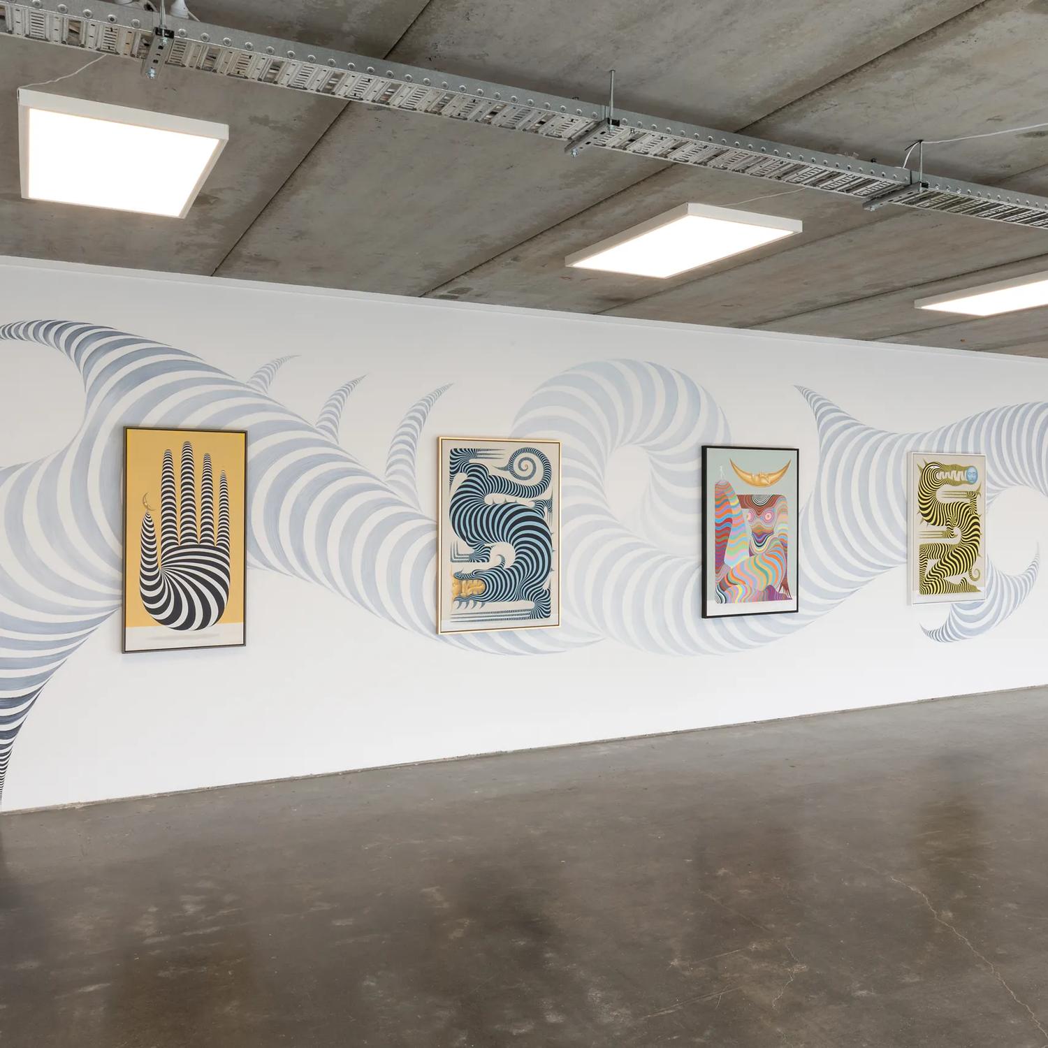 A large room wth white walls, a concrete floor and an exposed ceiling with a collection of contemporary prints on display.