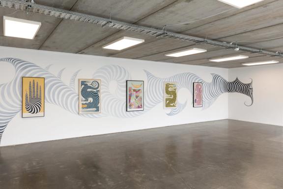 A large room wth white walls, a concrete floor and an exposed ceiling with a collection of contemporary prints on display.